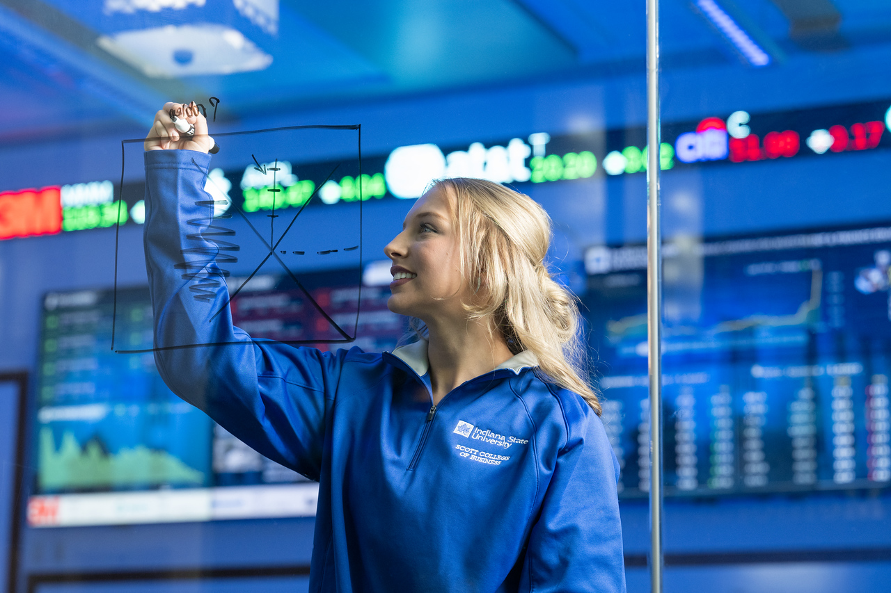 White female with blonde hair wearing a blue Indiana State University Scott College zip-up stands in front of large digital stock market displays with ticker symbols and charts, holding a marker and writing on the wall.