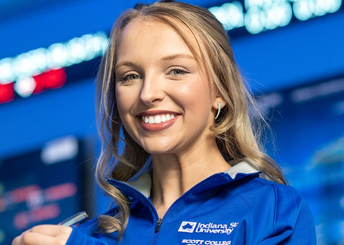 White female with blonde hair wearing a blue Indiana State University Scott College zip-up stands in front of large digital stock market displays with ticker symbols and charts.