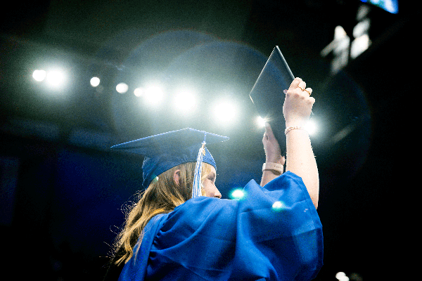 Graduate in a blue cap and gown seen from behind on a stage under bright lights, raising a diploma folder overhead during a commencement ceremony.