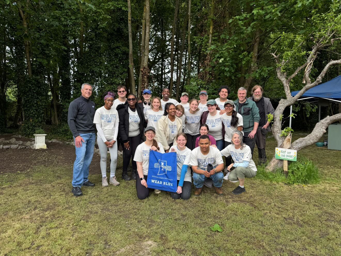 A group of people, some wearing "Wear Blue" shirts, stand and kneel on grass in a wooded area. One person holds a sign with the "Indiana State University: Wear Blue" logo. There's a small tent and a "Plant Sale" sign nearby.