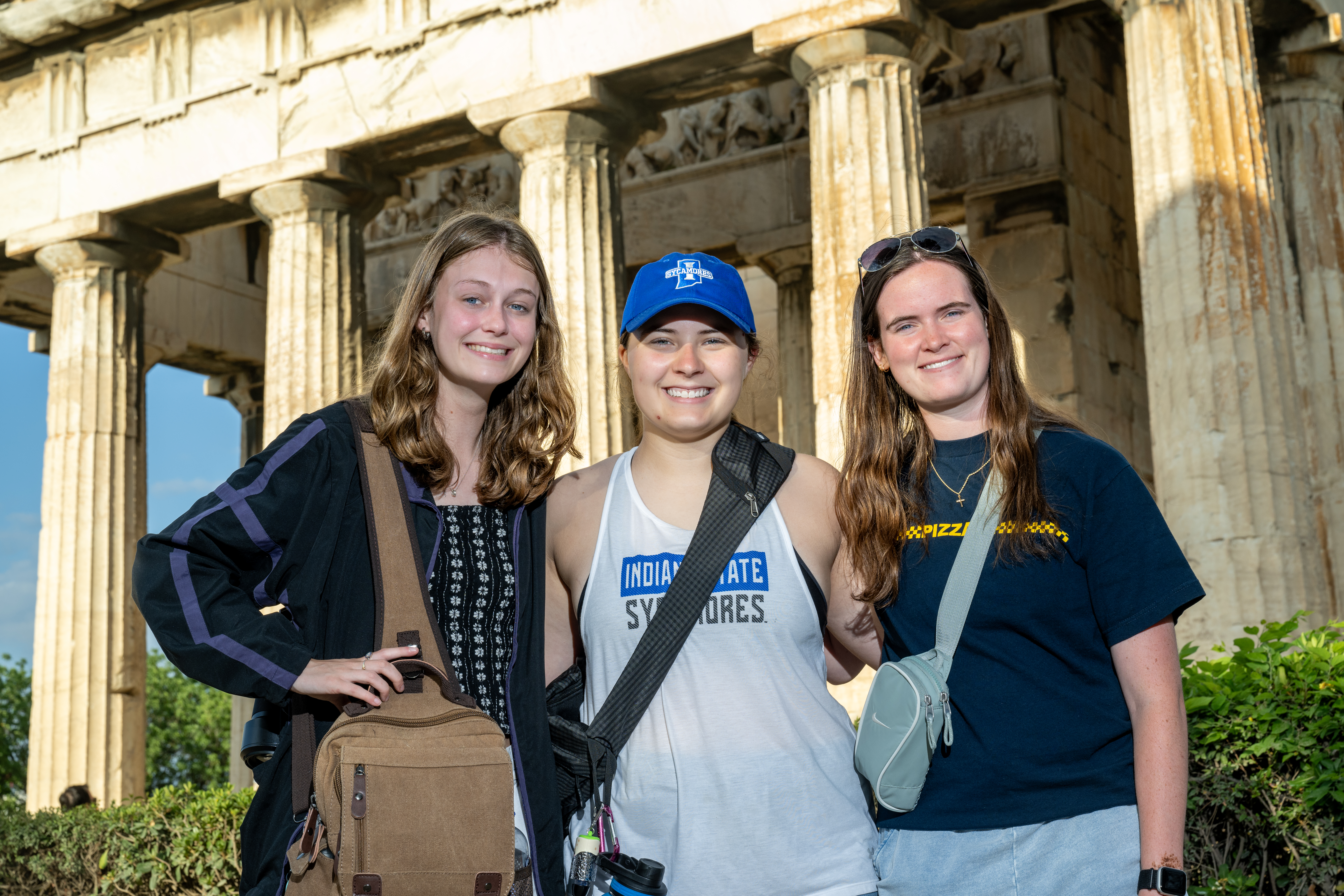 Three women stand together in front of a historic Greek stone building with tall columns, wearing casual clothing and carrying small bags, with the central person wearing an Indiana State Sycamores shirt and cap. Sunlight illuminates the structure behind them.