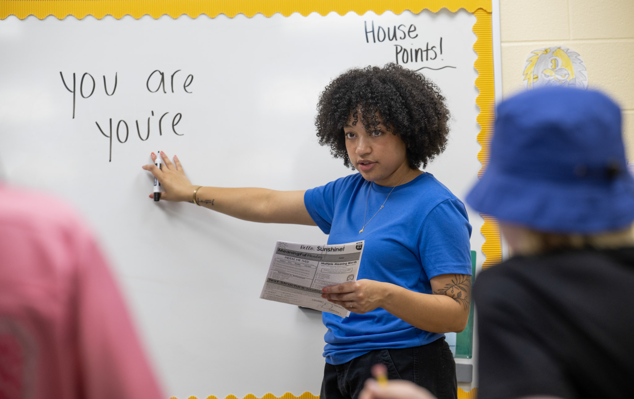 A student teacher stands at a whiteboard, pointing to the words "you are" and "you're" written on it. They hold a paper titled "Worksheet" and are wearing a blue shirt. The board is bordered with yellow paper, and "House Points!" is written in the corner. Two younger children are partially visible in the foreground.
