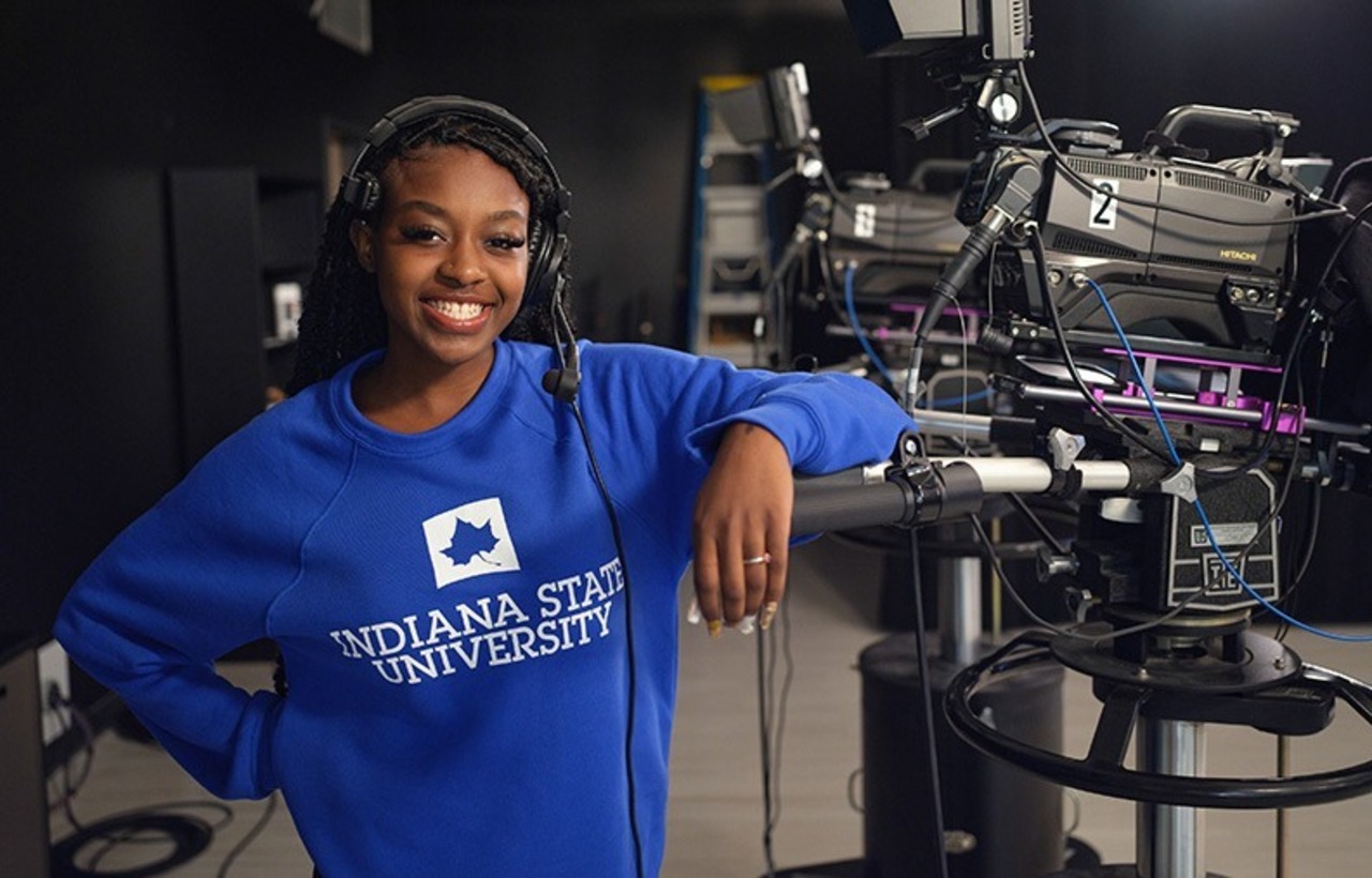 A person in a blue "Indiana State University" sweatshirt, wearing a headset, stands smiling in a television studio next to a large video camera.