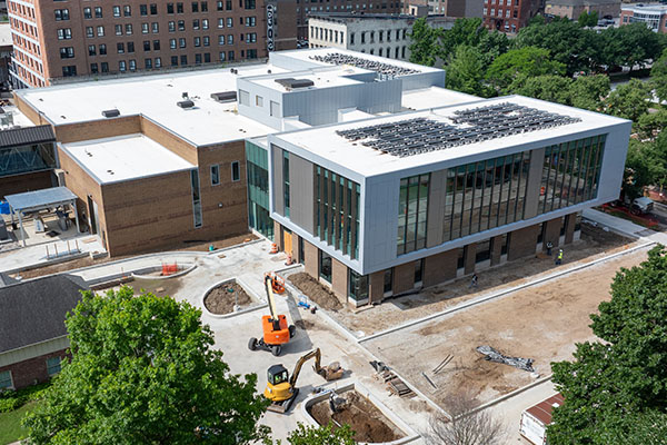 Aerial shot of a glass, brick, and cement building standing amidst a construction site with patches of dirt and open foundations, yellow and orange construction equipment, green trees, and other buildings visible around and behind it. 