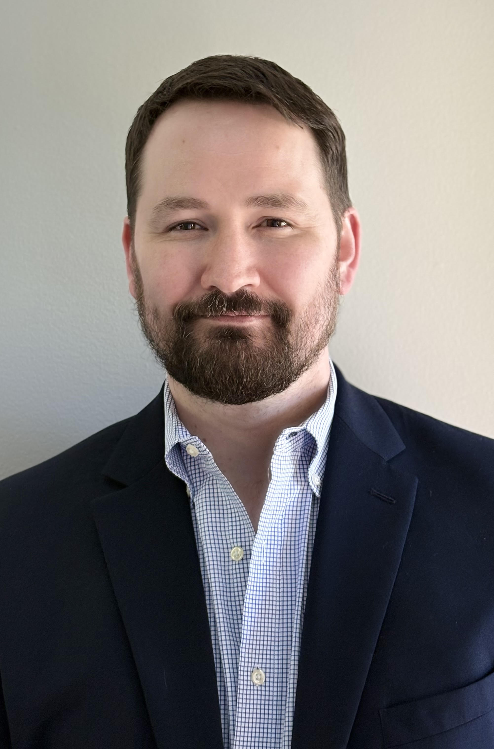 Professional headshot of an adult with short dark hair and a neatly trimmed beard, facing the camera with a slight smile. They are wearing a navy blazer over a light blue checkered button-down shirt. The photo is framed from the chest up against a plain light-colored wall, with soft, even lighting and a clean, business-professional appearance.