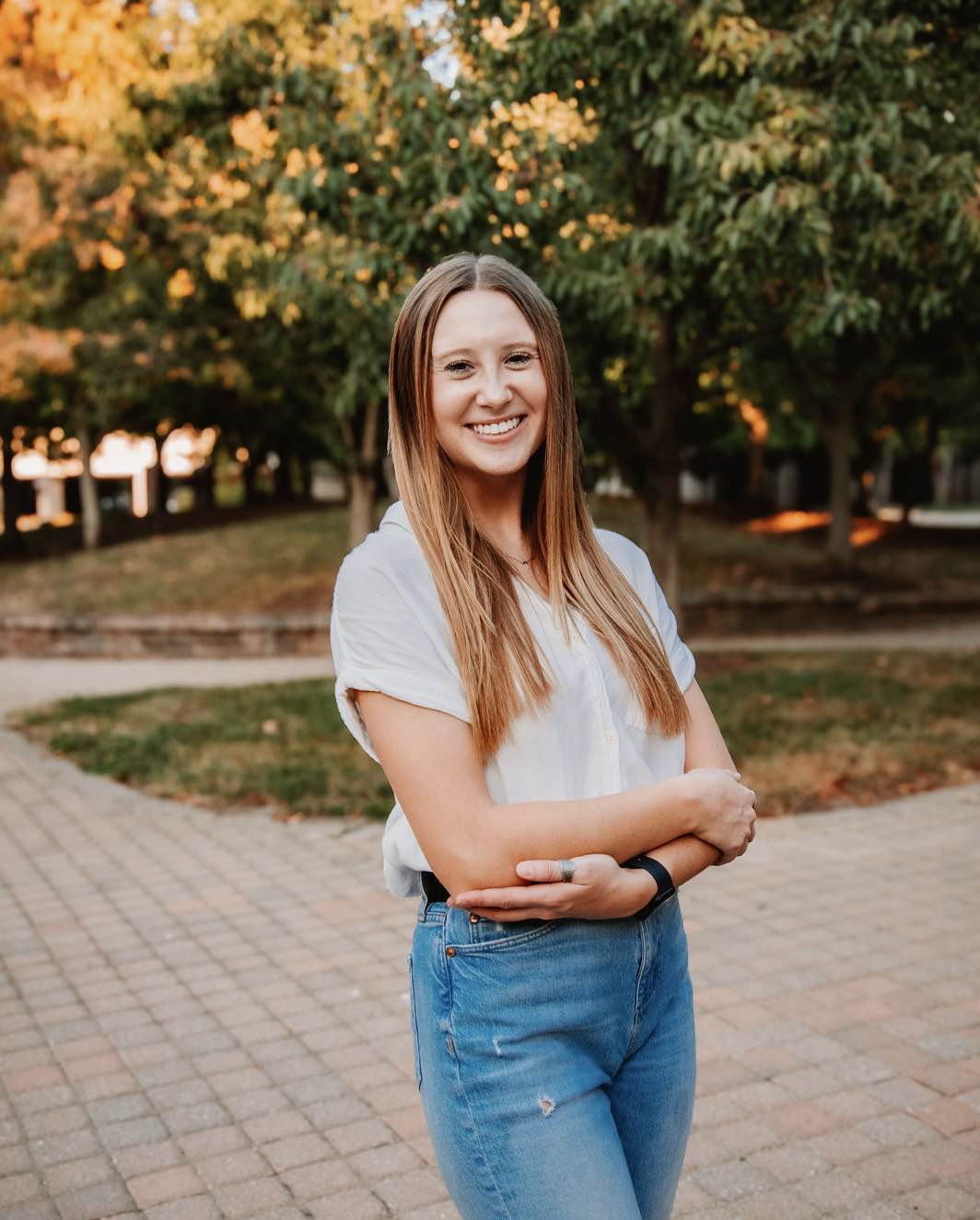 Smiling woman standing on a paved walkway with arms crossed, wearing a light short-sleeve shirt and jeans, with trees and campus landscaping in the background.
