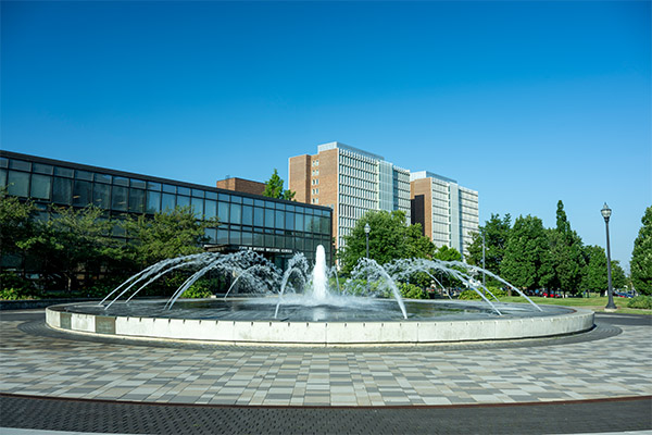 Circular fountain with multiple arcing water jets centered in a paved plaza on the Indiana State University campus. Low, modern academic buildings and taller residence hall-style buildings stand behind the fountain, with green trees and a clear blue sky in the background.