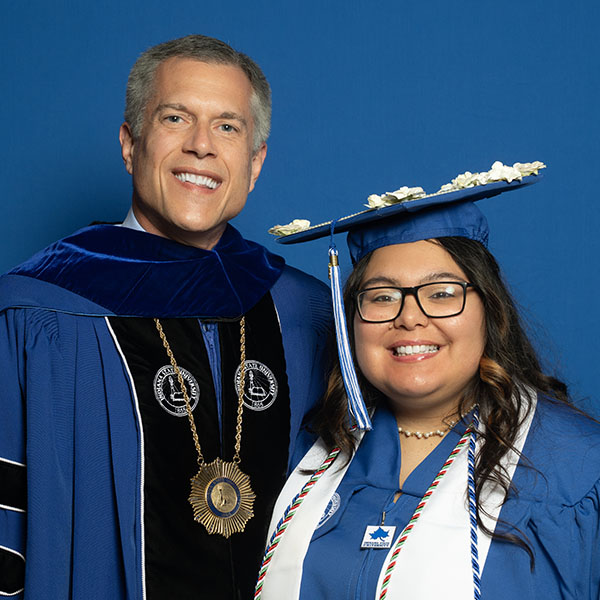 Graduate in cap and gown stands beside Indiana State University President Mike Godard, who is dressed in academic regalia. Both face the camera against a blue studio background. The graduate wears glasses, a decorated mortarboard, honor cords, and a stole, while President Godard wears a ceremonial medallion and gown.