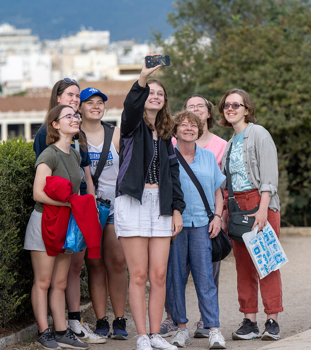 Group of people standing on a path beside a hedge, gathered close together while one person holds up a phone to take a selfie, with buildings and trees in the background.