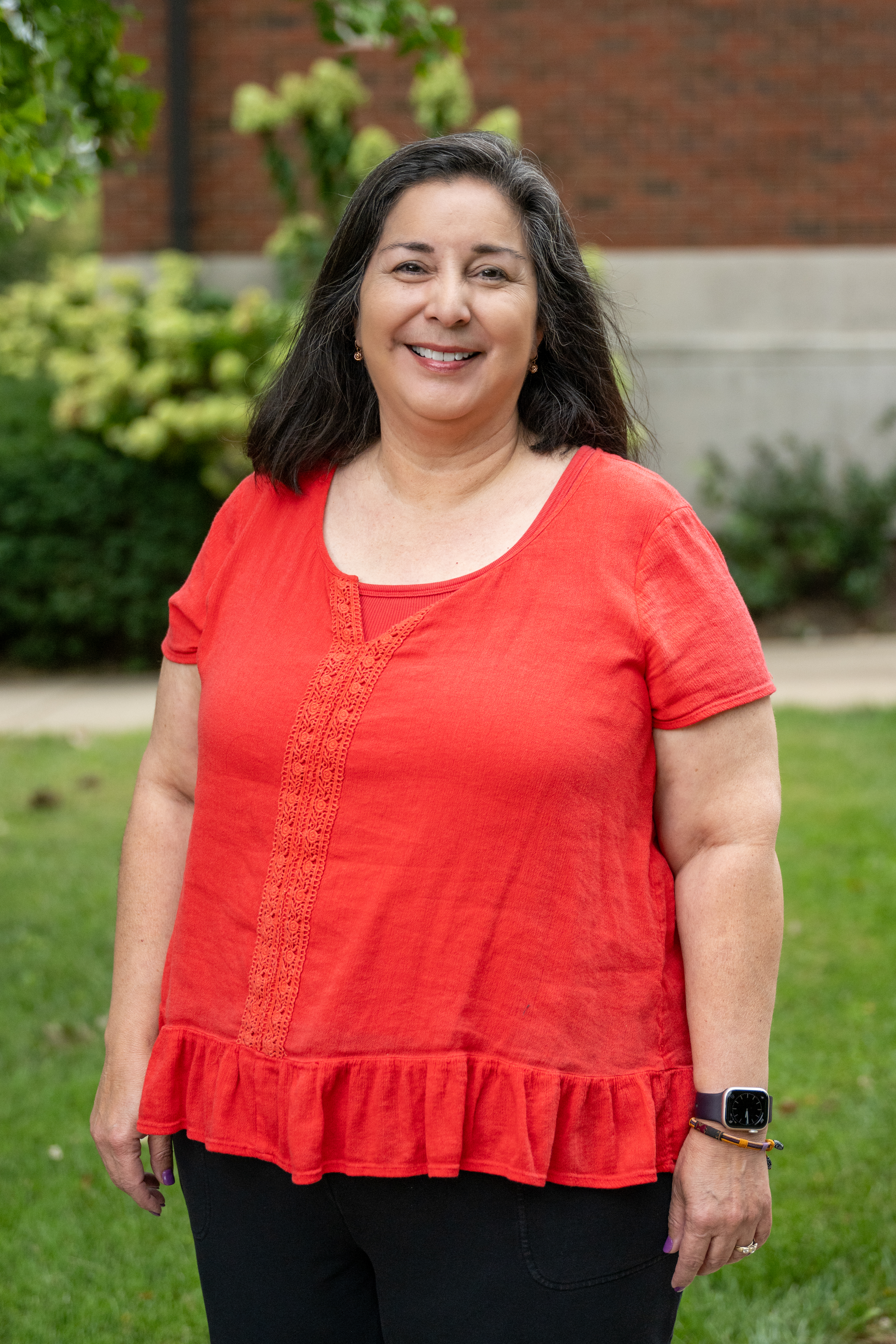 A smiling woman with shoulder-length dark hair stands outdoors on a grassy lawn, wearing a bright coral-red short-sleeve blouse, black pants, and a smartwatch, with a brick building and greenery blurred in the background.