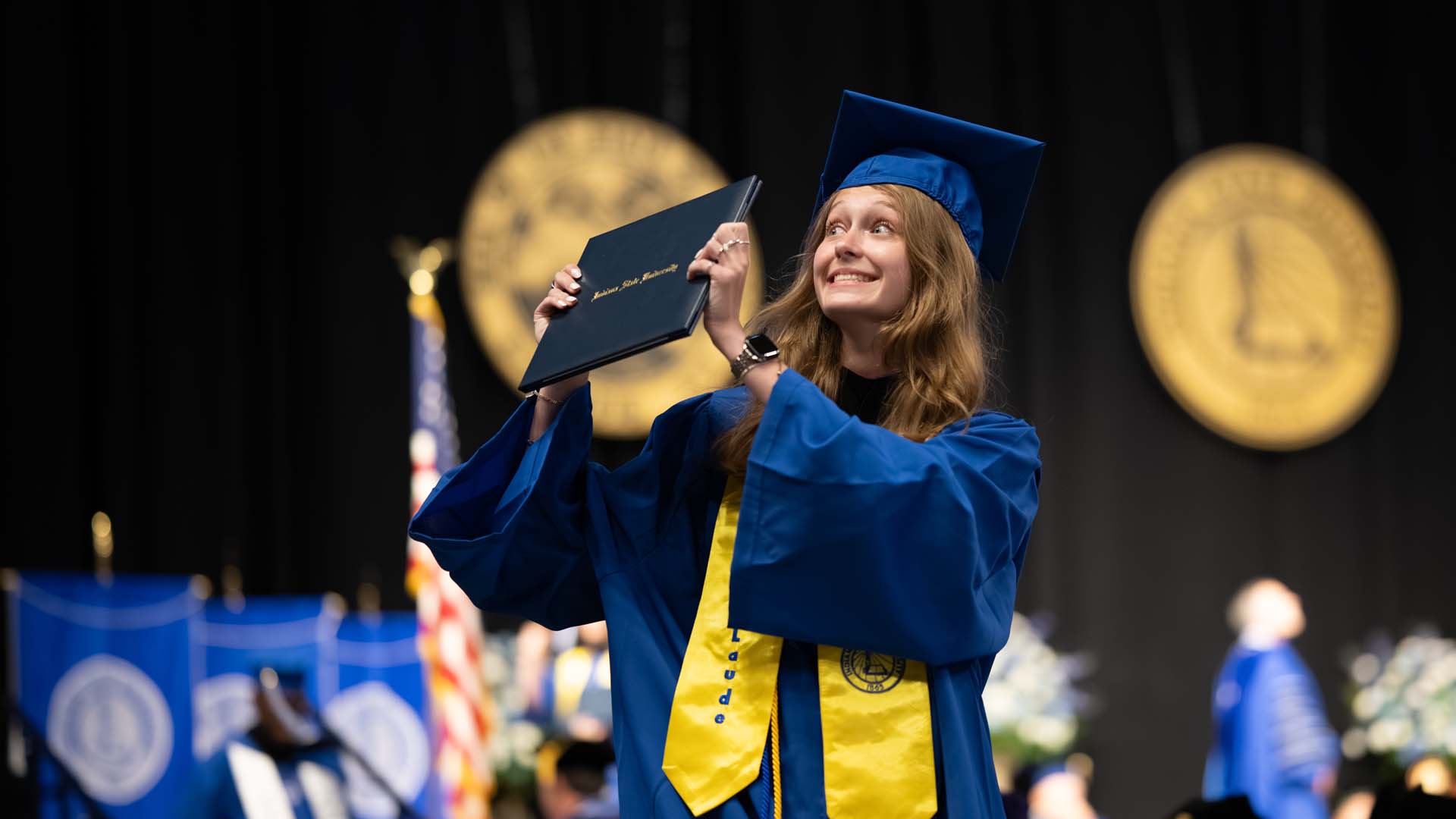 Smiling young woman in blue cap and gown and a yellow stole holds up a diploma on a stage during a commencement ceremony, with blurred university seals, flag, and figures in the background.