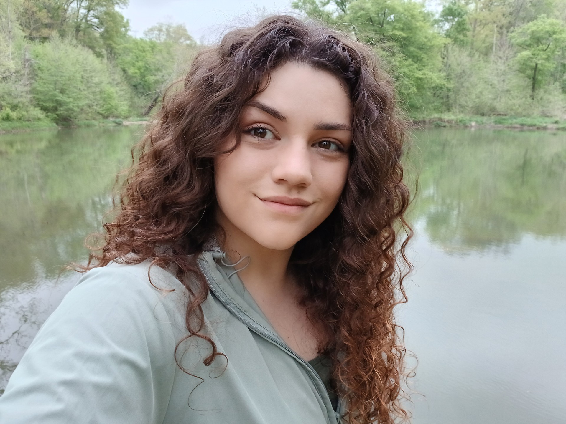Close-up outdoor portrait of a person with long, curly dark brown hair taking a selfie beside a calm body of water. They are wearing a light gray-green zip-up jacket and looking at the camera with a slight smile. Trees and greenery line the opposite shore, and the water reflects the surrounding landscape under an overcast sky.