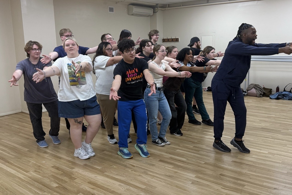 Group of about 15–16 young adults in a rehearsal room with wood floors stand in staggered rows, extending both arms forward in a synchronized movement while following an instructor at the front right of the group.