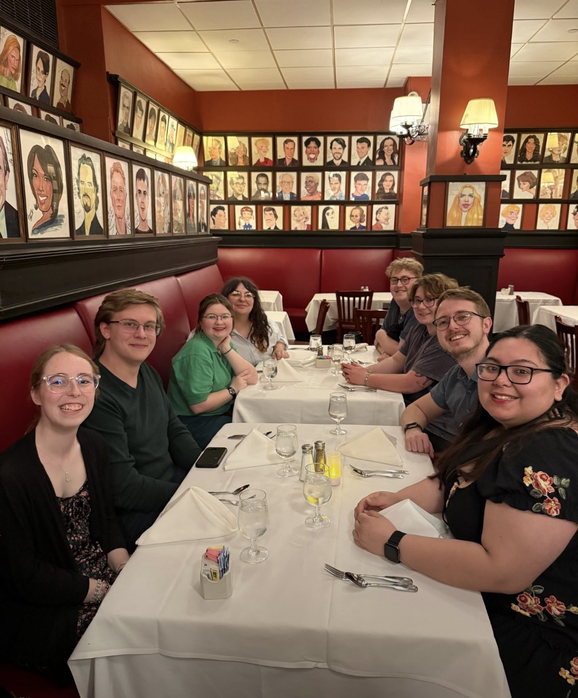 Group of eight young adults seated around a white tablecloth in a restaurant with red walls and booth seating, posing for a photo. The walls are lined with framed caricature portraits, and the table is set with folded napkins, water glasses, and silverware.