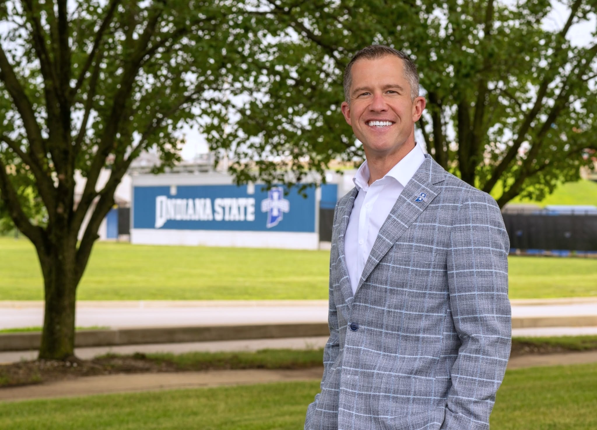 A person in a gray plaid suit stands smiling outdoors with trees and grass in the background. A building with signage that reads "Indiana State" is visible in the distance.