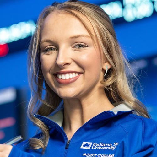 Close-up portrait of a person wearing a blue Indiana State University quarter-zip jacket with “Scott College” text visible on the chest, smiling toward the camera against a softly blurred blue background with lights.