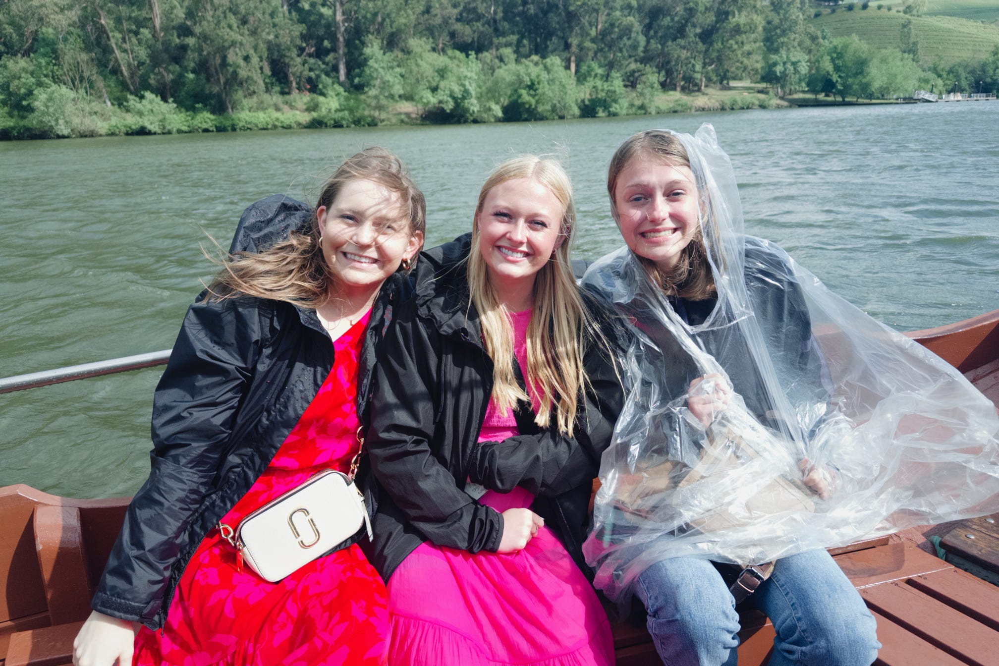 Three young women sit close together on a boat wearing jackets, with a lake and tree-lined shoreline behind them. 
