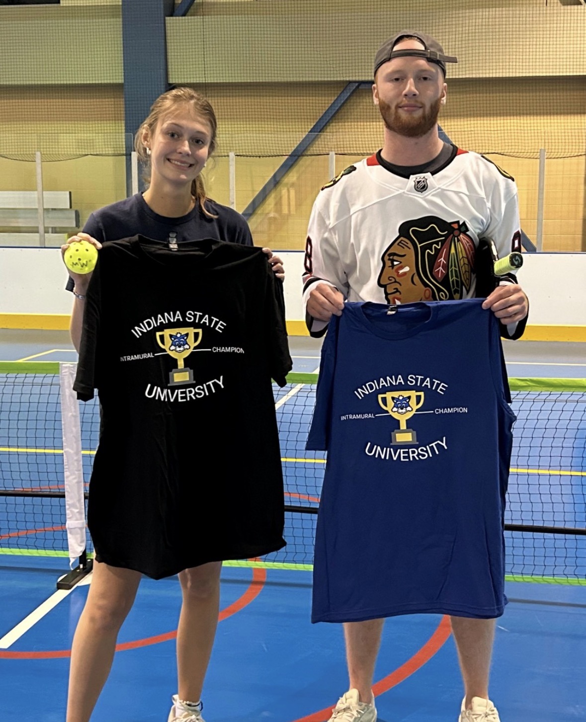 Two people stand on an indoor sports court holding Indiana State University Intramural Champion T-shirts, one black and one blue, with a tennis net and court lines visible in the background.