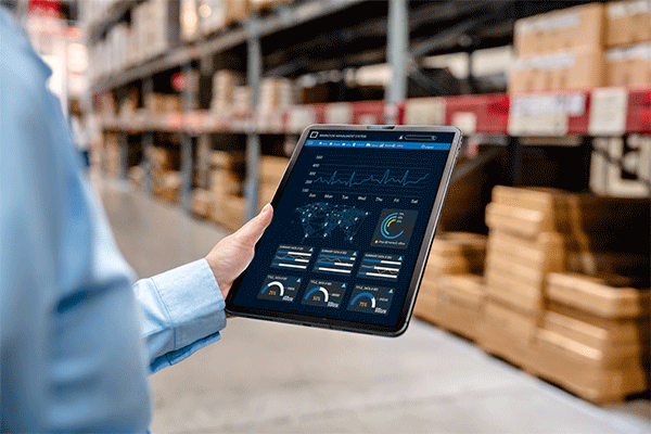 Person’s hand holding a tablet displaying data dashboards in a warehouse aisle with shelves of stacked boxes in the background.