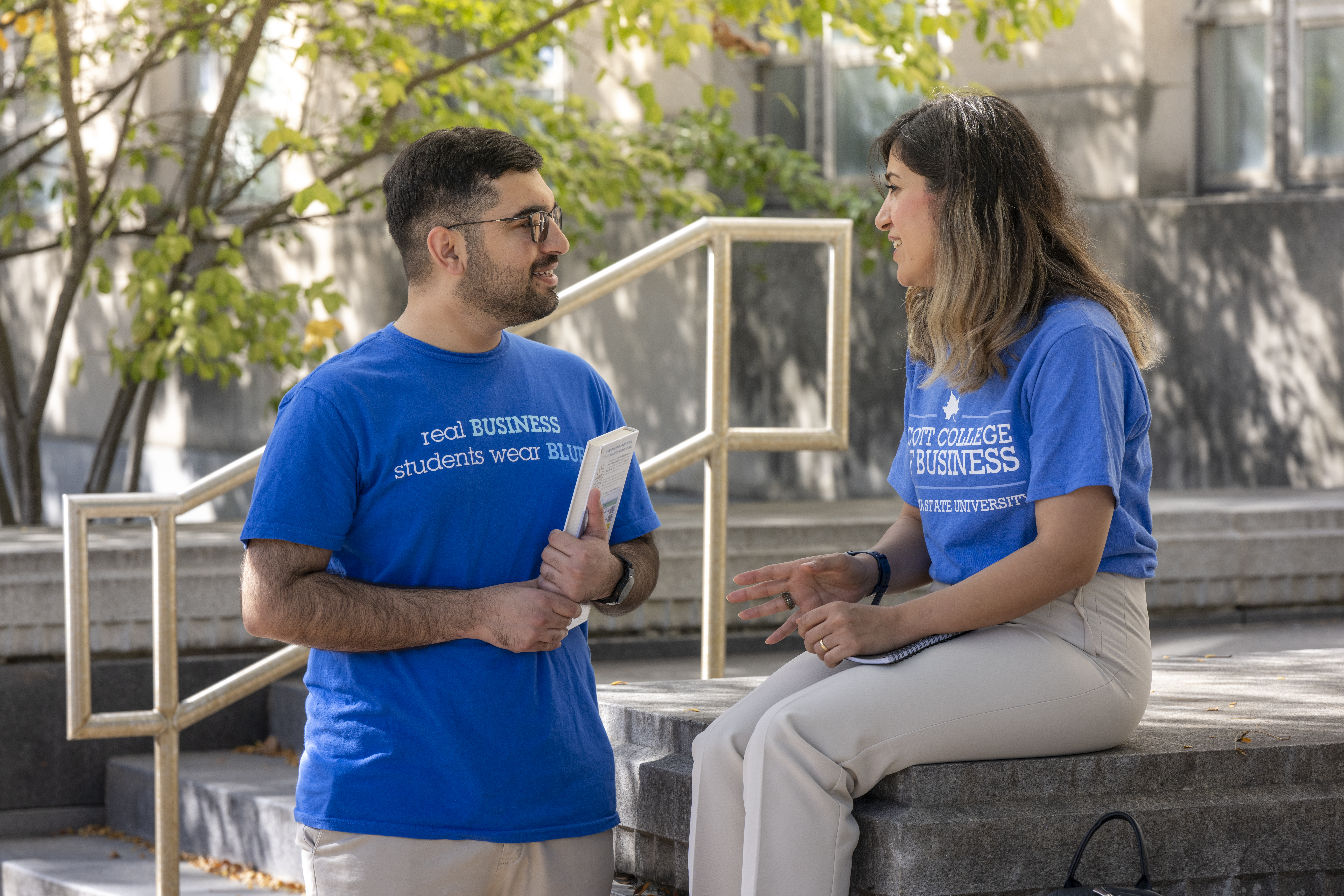 Two people in blue shirts converse outdoors on a sunny day. One person's shirt reads, "Real Business Students Wear Blue." The other person's shirt says, "Scott College of Business." They are near steps and surrounded by trees. One holds a notebook, and the other is seated on a stone surface.