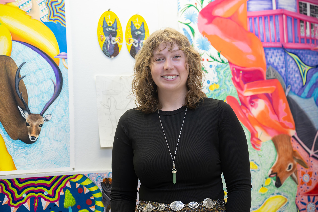 Female student with short curly brown hair standing in an art studio surrounded by brightly colored paintings and mixed-media artwork featuring animals, abstract patterns, and surreal imagery.