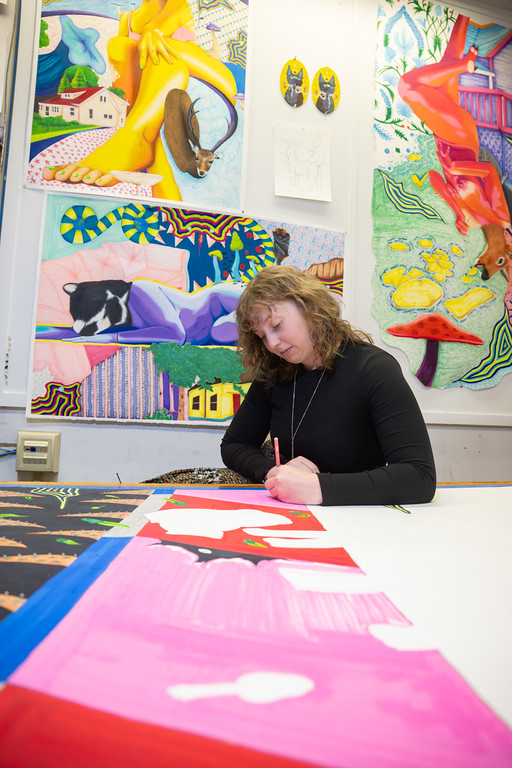 Female student with short curly brown hair sketching at a large worktable in an art studio, surrounded by colorful paintings featuring abstract patterns, animals, and surreal imagery displayed on the walls.