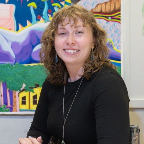 A female student with short curly brown hair seated at a worktable in an art studio, with colorful paintings and mixed-media artwork displayed on the walls behind her.