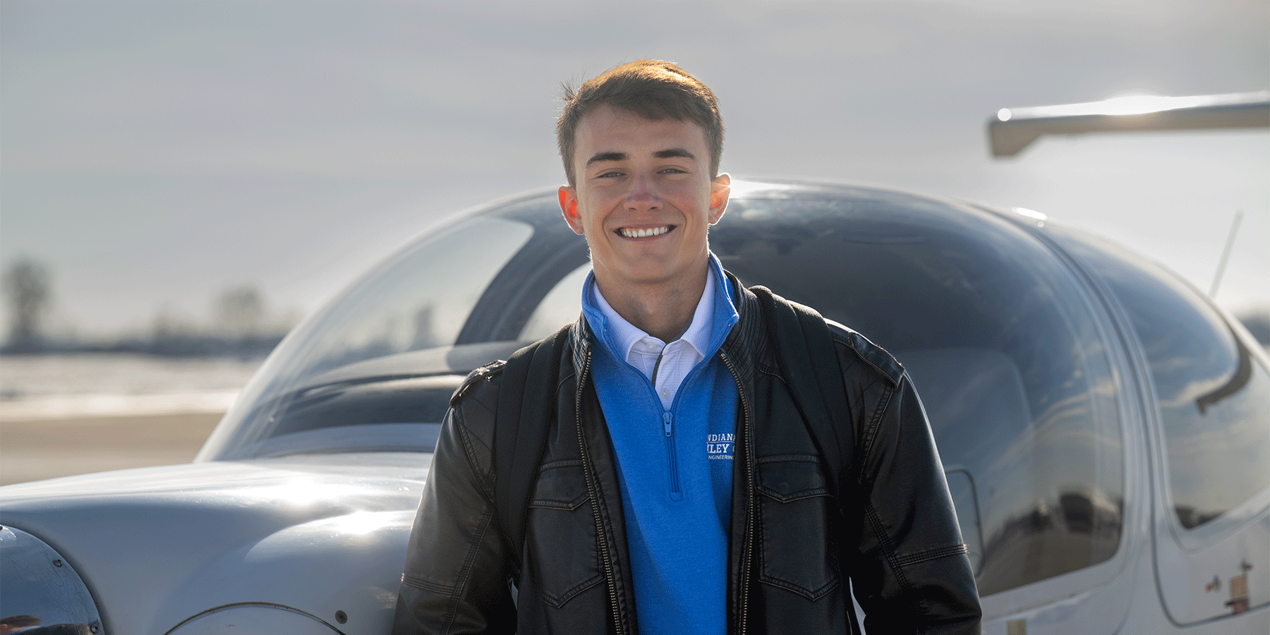 Male student with a backpack stands in front of a small aircraft on an airport tarmac, smiling and looking toward the camera.