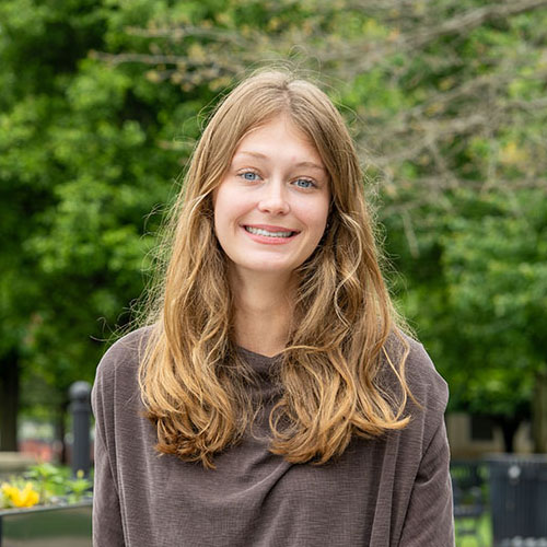 Person with long hair wearing a brown shirt, standing outdoors on a campus walkway with trees and flowers in the background.