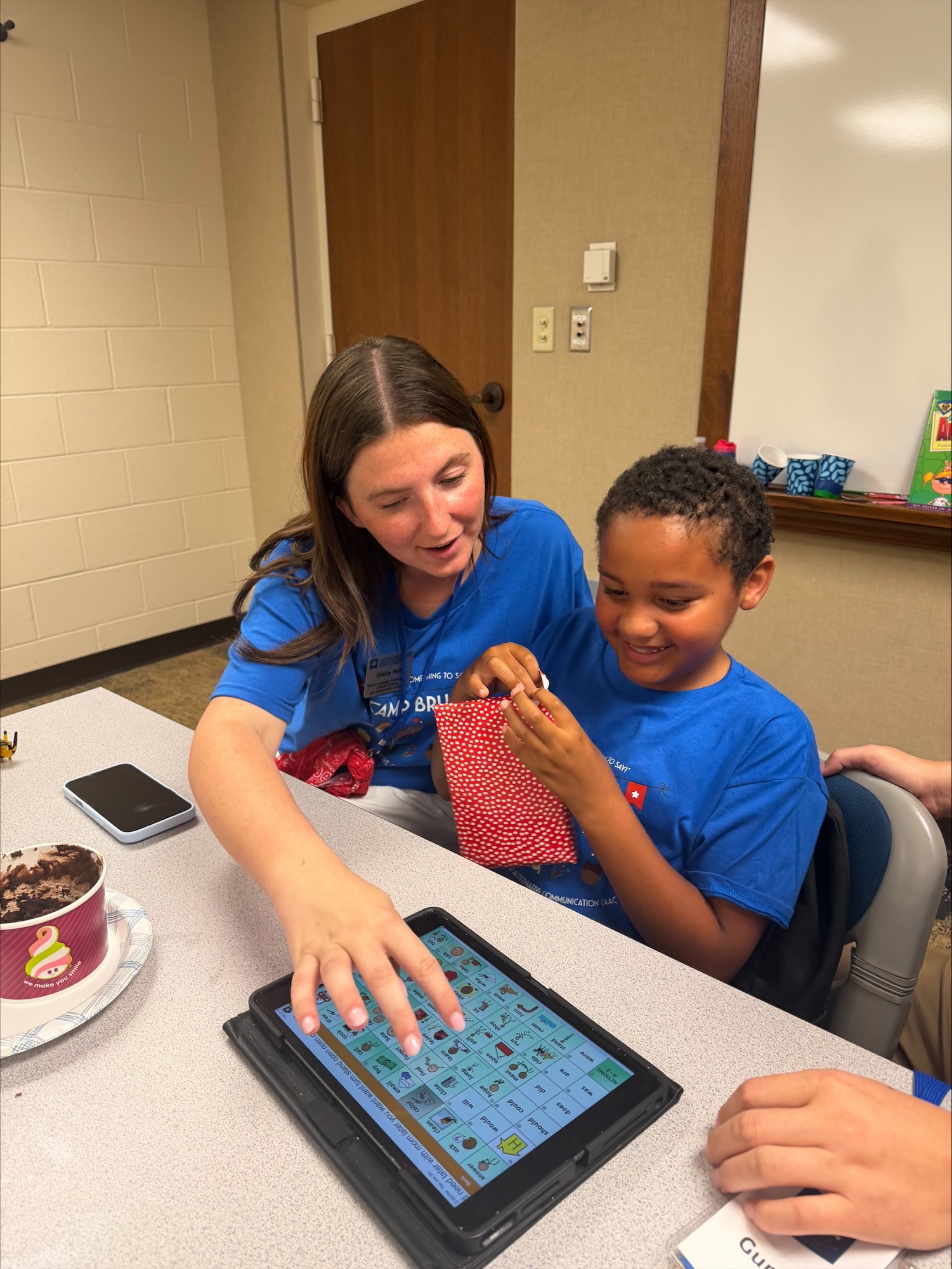 A student assists a young child with an augmentative and alternative communication (AAC) device.