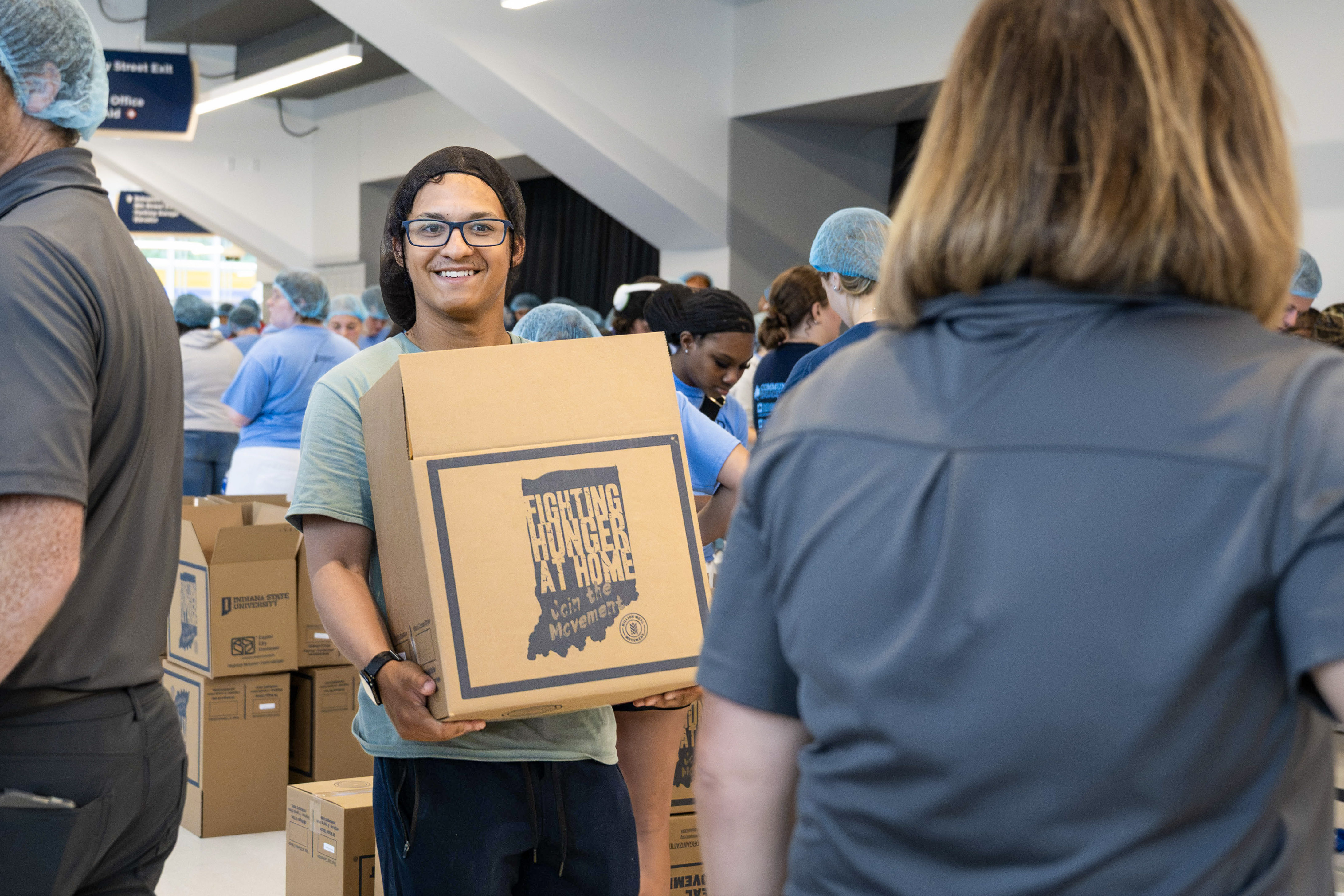 A person carrying a box labeled "Fighting Hunger at Home" at a community event. People in the background are wearing hair nets and sorting boxes.