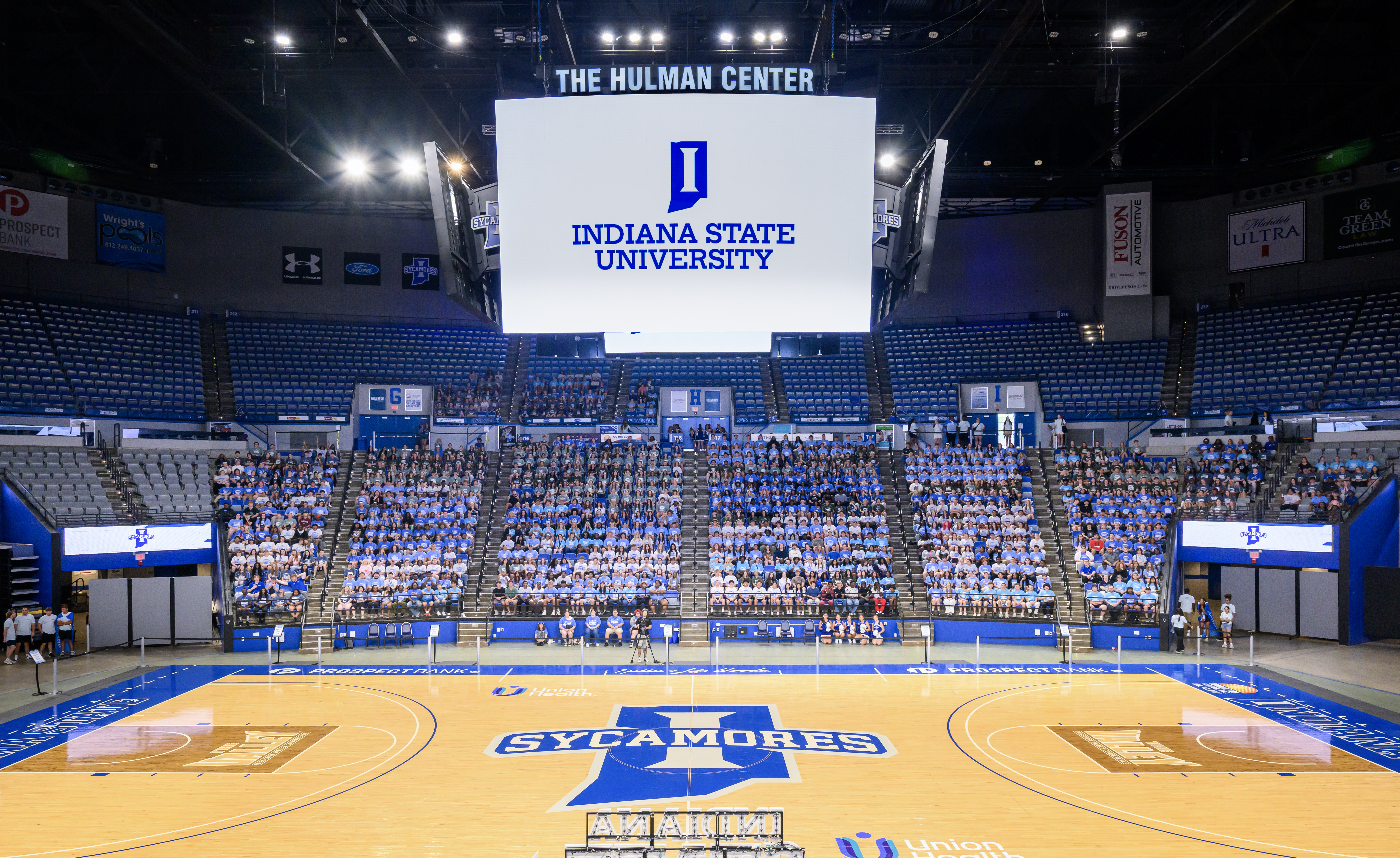 The image shows the interior of the Hulman Center at Indiana State University. It features a large basketball court with the Sycamores logo at center court. A big screen above displays the university's logo and name. The seating area is filled with people, most wearing white shirts.