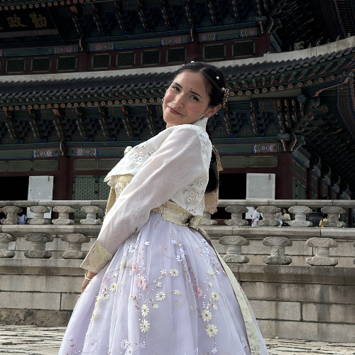 Person wearing a traditional hanbok stands on stone steps in front of an ornate historic building with decorative wooden architecture. The outfit features a light-colored embroidered skirt and jacket, and the building’s detailed roofline fills the background.