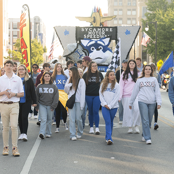A large group of students walks down a city street during a parade, led by participants wearing Greek organization sweatshirts. Behind the group is a float featuring the Sycamore Sam mascot and a sign reading “Sycamore Motor Speedway,” with flags and downtown buildings visible in the background.
