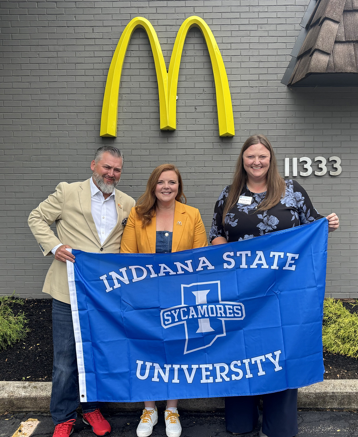 An image features three individuals holding a large, blue "Indiana State University Sycamores" flag in front of a grey brick building adorned with a prominent yellow McDonald's logo.