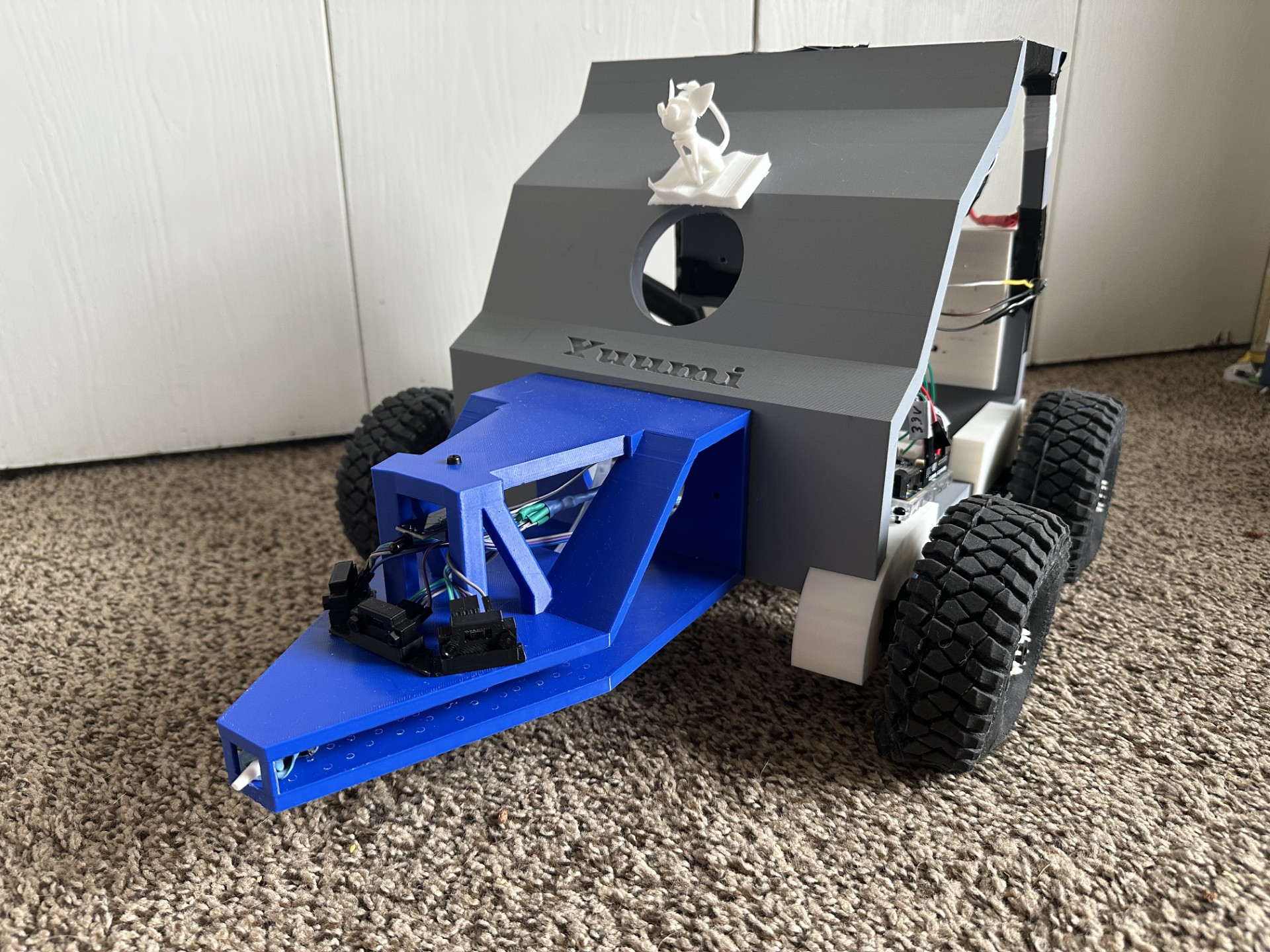 Four-wheeled robotic vehicle with large tires and a gray 3D-printed body sits on a carpeted floor. A blue front-mounted attachment with exposed wiring extends from the front, and a small 3D-printed figure is mounted on top.