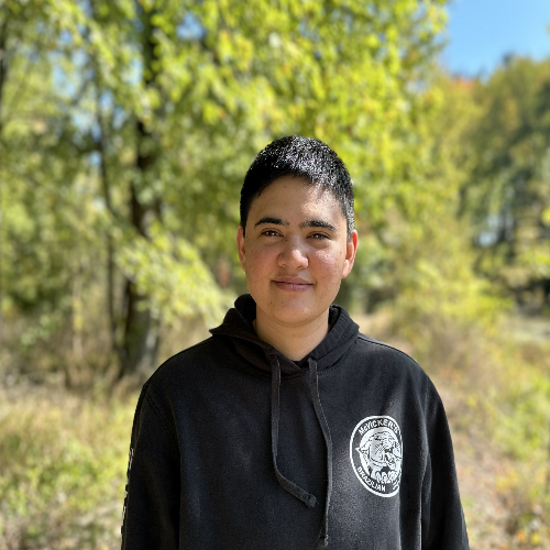 Person with short dark hair wearing a black hoodie stands outdoors on a sunny day, with green trees and a softly blurred wooded background.