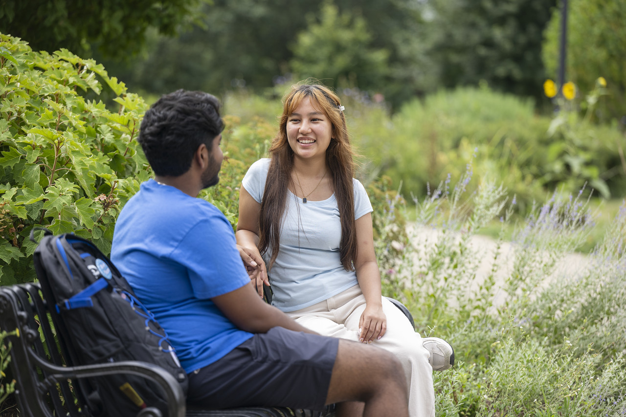 Two students sit on a bench along a landscaped campus path, talking outdoors among trees, shrubs, and flowering plants. A backpack rests on the bench beside one student.