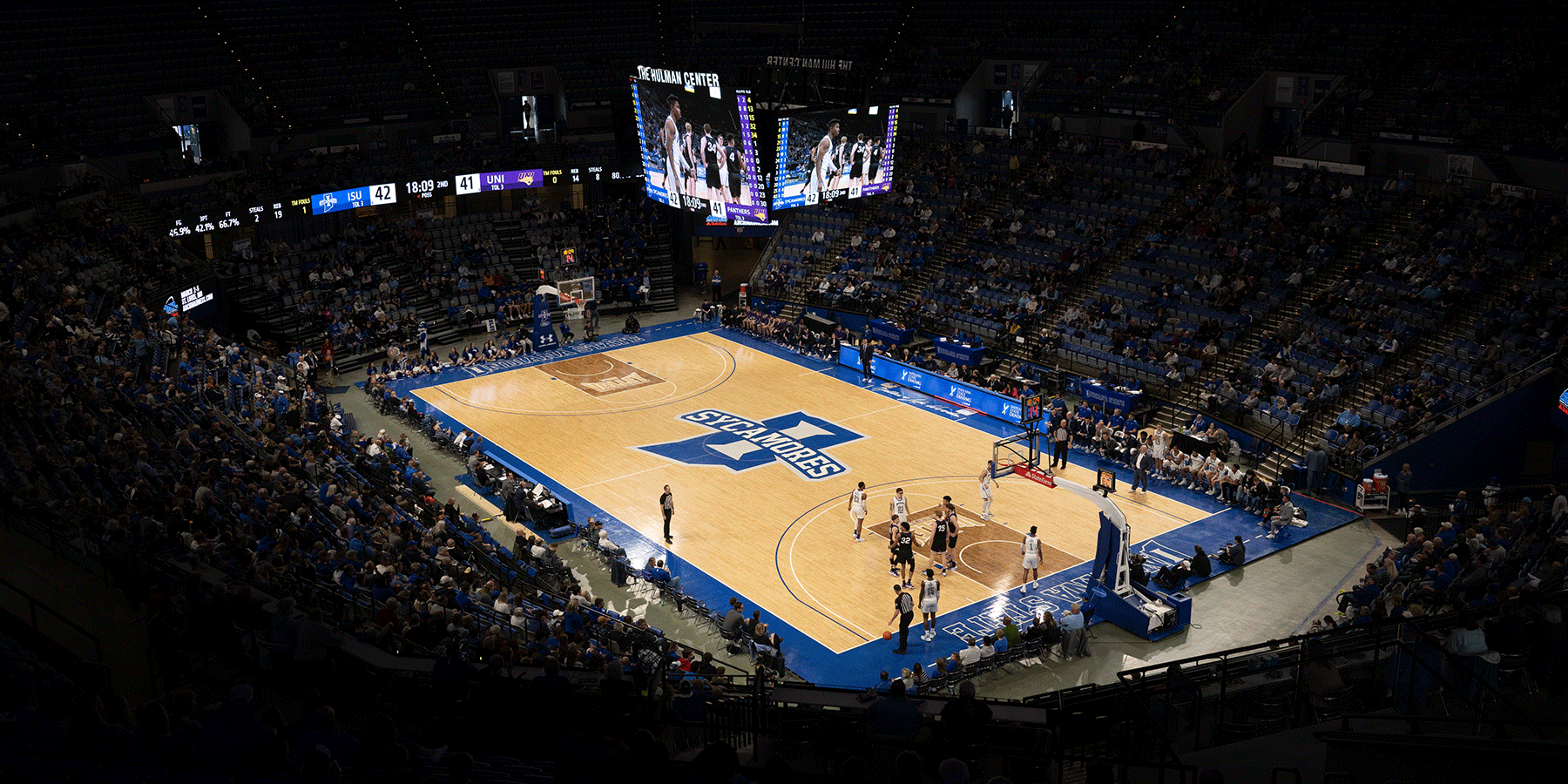 Wide view of an indoor basketball arena during a game, showing players on the court, spectators seated around the venue, and a large center-hung scoreboard above the floor. The court features a large “Sycamores” logo at center court, and the scoreboard displays a close score between Indiana State and UNI in the second half.