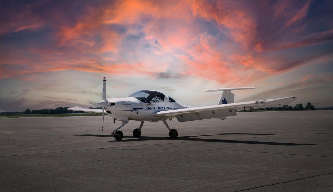 An airplane branded with "Indiana State University" is parked on a runway. The sky in the background features a vibrant sunset with hues of orange, pink, and purple.