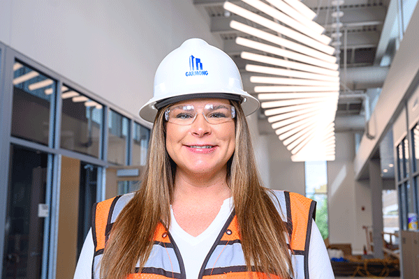 Female wearing a white hard hat, clear safety glasses, and an orange reflective safety vest stands inside a building under construction. Large floor-to-ceiling windows and green trees are visible in the background.