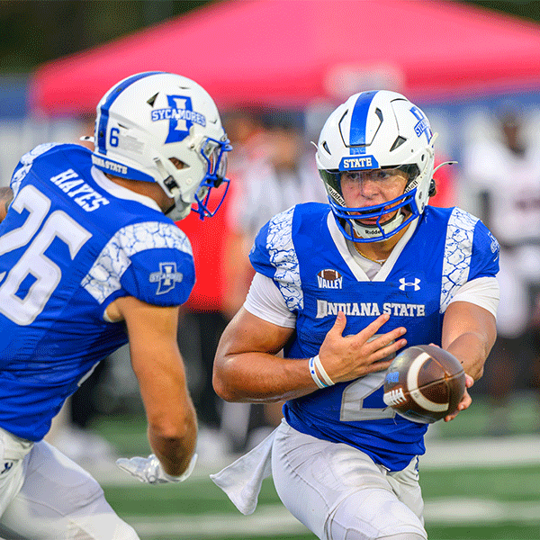 Indiana State football player carries the ball during a running play while a teammate moves ahead to block. Both players wear blue and white uniforms and helmets, with a blurred red canopy visible in the background.