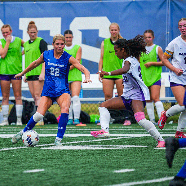 Soccer player in a blue Indiana State uniform dribbles the ball while a defender in a white uniform attempts to challenge for possession on a turf field. Players in bright green training pinnies watch from the sideline in the background.