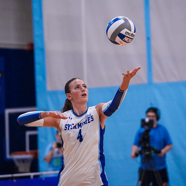 Volleyball player in a white Indiana State uniform tosses the ball into the air to serve during an indoor match, with the ball suspended above an outstretched hand.