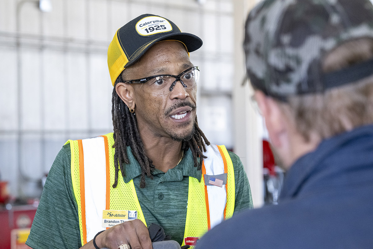 A man wearing safety glasses, a high-visibility safety vest, and a Caterpillar-branded cap speaks with another person in an industrial workshop or maintenance facility. The second person is out of focus in the foreground. Equipment and work areas are visible in the background.