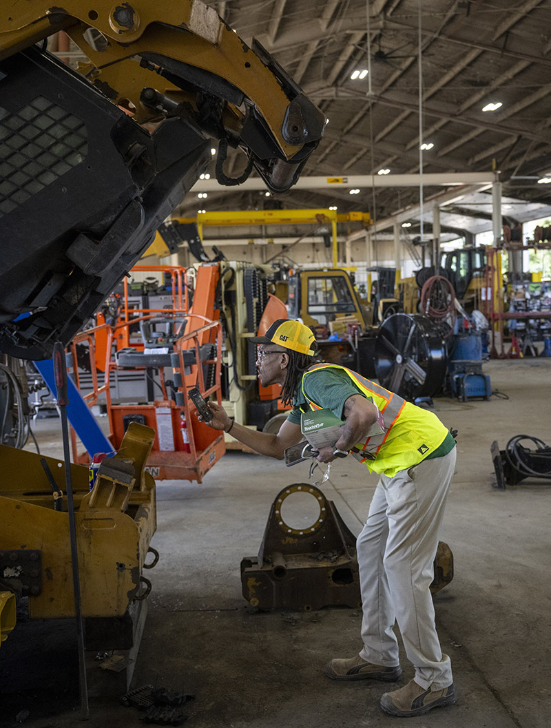 A person wearing a high-visibility safety vest, work boots, and a yellow CAT cap inspects a large piece of construction equipment inside a maintenance workshop. The person holds a handheld device while examining the raised machine. Additional heavy equipment, tools, lifts, and industrial work areas are visible throughout the facility.