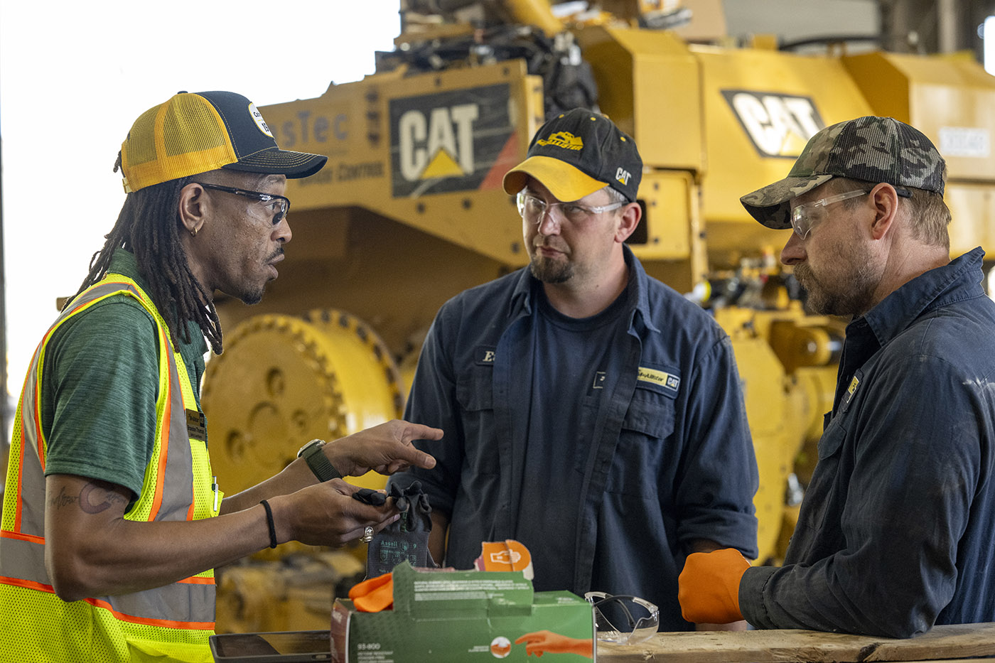 Three people wearing safety glasses stand beside large CAT equipment in a maintenance workshop. One person in a high-visibility safety vest holds a pair of work gloves while speaking with two people in work uniforms. Boxes of protective gloves and safety gear are visible on a work surface in the foreground.