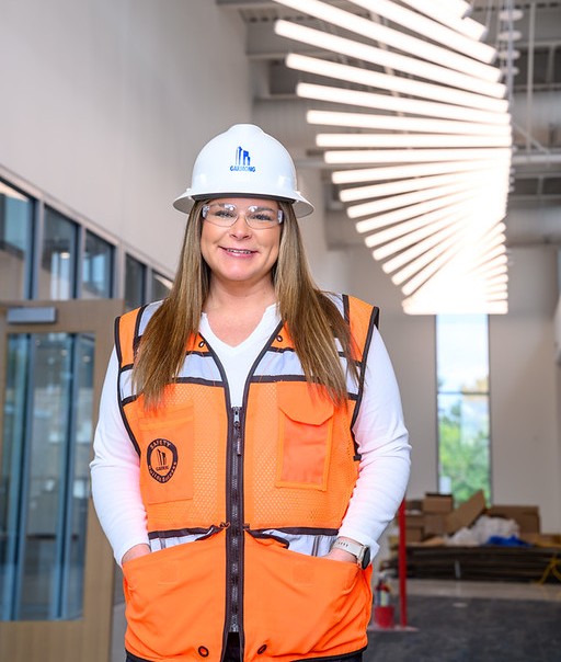 Female wearing a white hard hat, safety glasses, and an orange reflective safety vest stands inside a modern building under construction. Decorative linear light fixtures hang from the ceiling overhead, with glass-walled rooms and construction materials visible in the background.