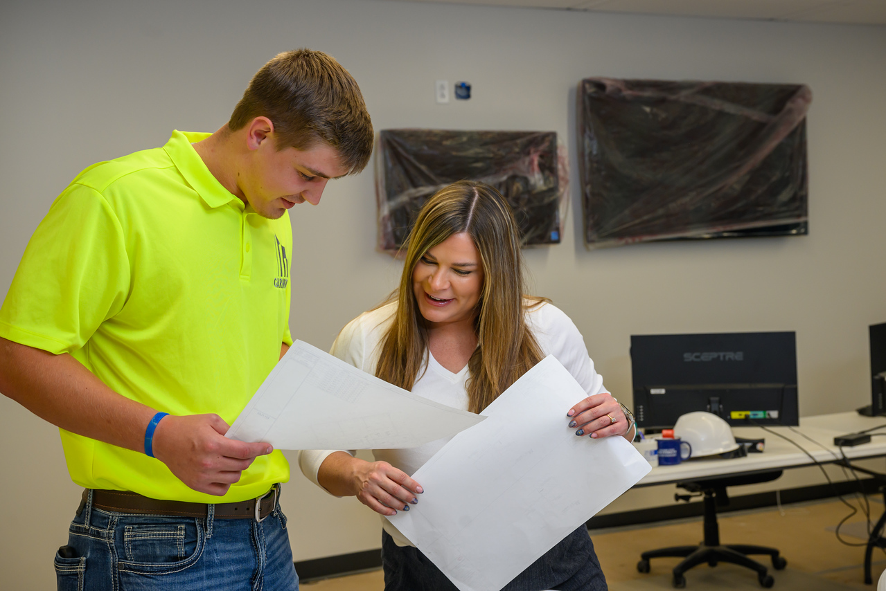 Two people review large construction plans in an office setting. One person wearing a bright safety shirt and another holding blueprints stand beside desks with computer monitors, while wall-mounted screens and construction equipment are visible in the background. 