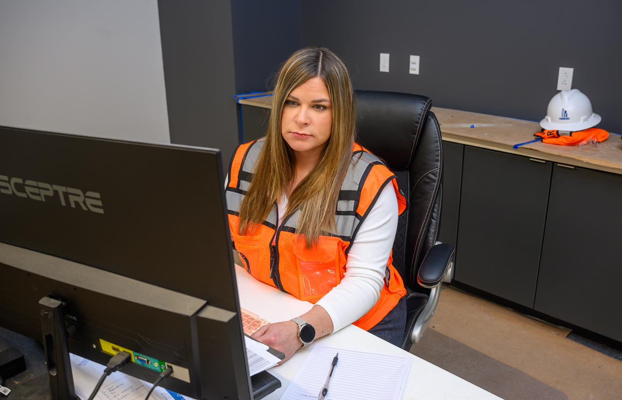 Female wearing an orange reflective safety vest sits at a desk working on a computer in an office environment. A hard hat and safety vest rest on a countertop behind the workstation, and a notebook and pen are visible on the desk.